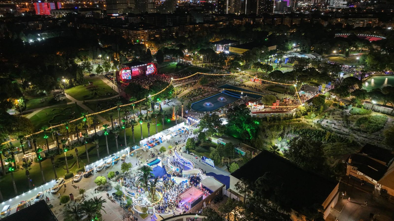 95. İzmir Enternasyonal Fuarı'nda Kültürpark'ın gece havadan görünümü; konser sahnesi önündeki kalabalık, aydınlatılmış havuzlar, palmiyeli aks ve stant sıraları